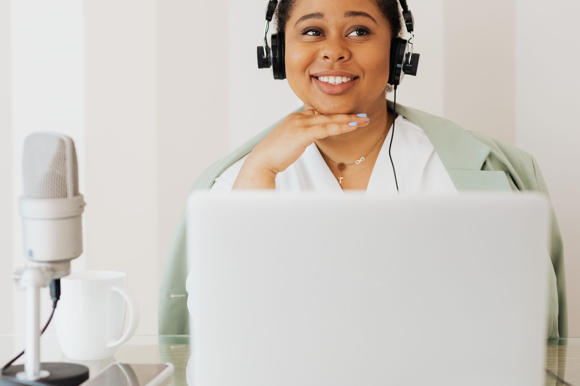 Una donna sorridente con le cuffie, seduta davanti a un portatile e a un microfono sulla scrivania di casa